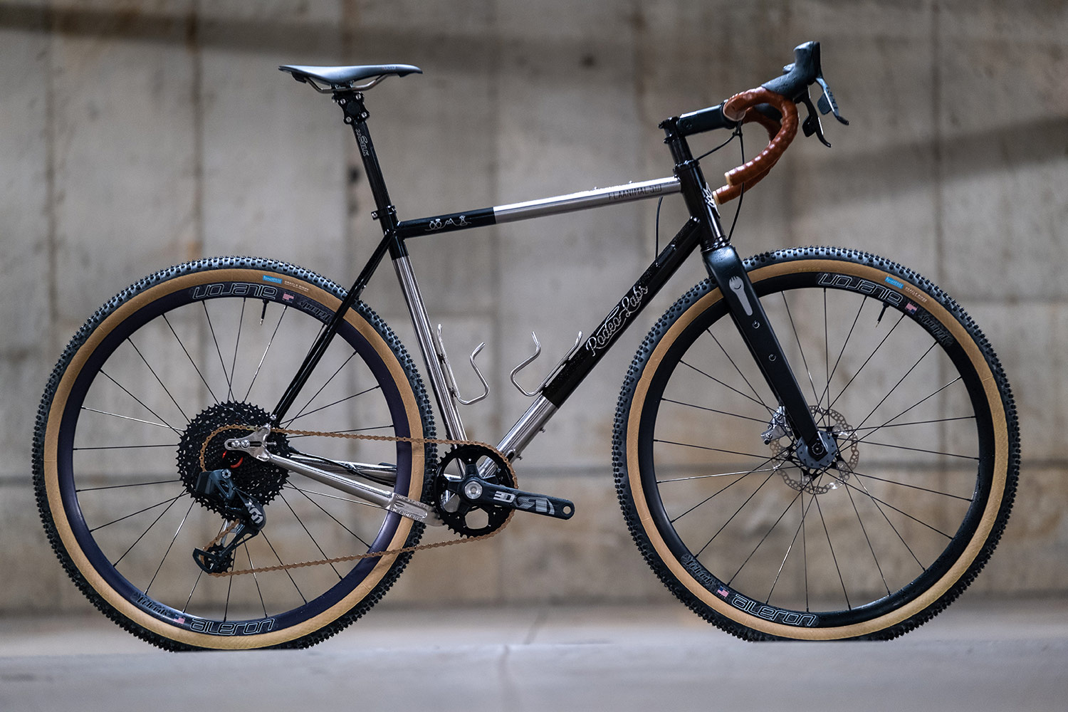 Black custom cerakote Rodeo Labs Flaanimal raw brushed titanium gravel bike with brown bar tape and tan wall tires.