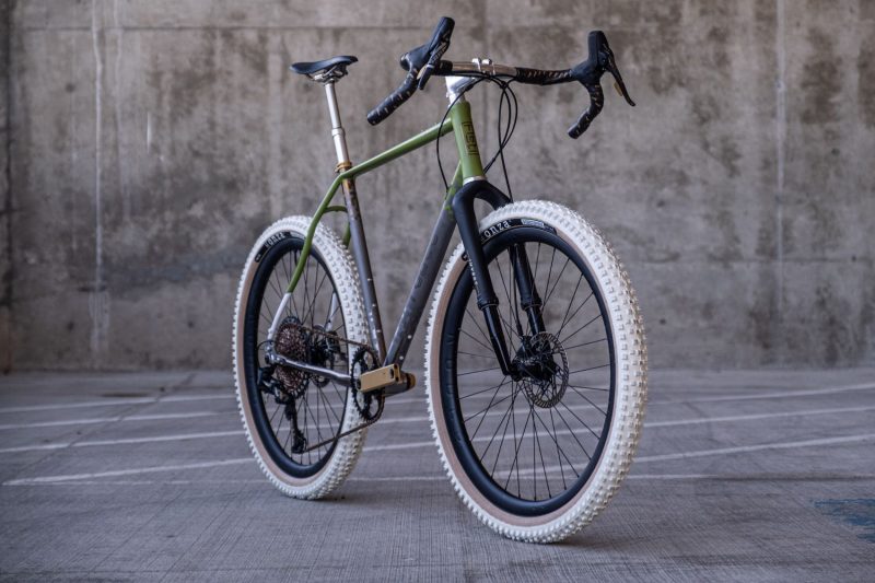 Rodeo Labs Flaanimal 6 Titanium gravel bike with a Cane Creek Invert suspension gravel fork and a silver PNW dropper post. This bicycle is custom cerakoted green and white, with gold anodized titanium logos and digital fade pattern. The wheels are Rodeo 3.0 carbon gravel wheels wrapped in white Onza Porcupine tires.